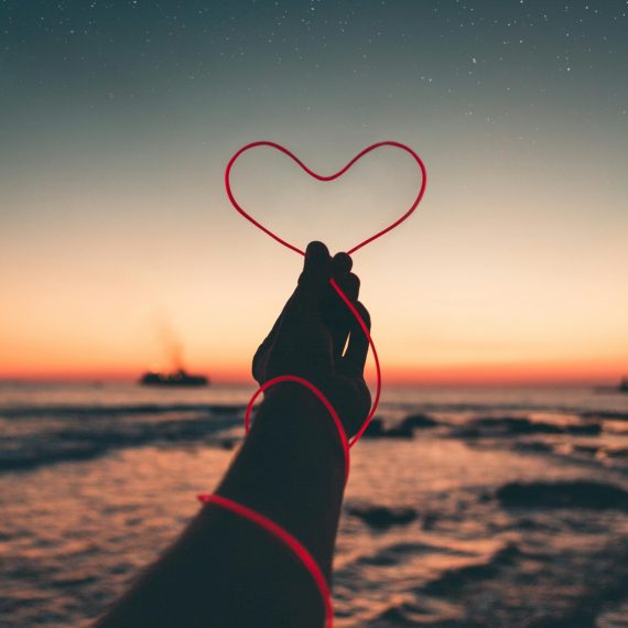 Silhouette of hand holding heart-shaped string at sunset by the sea.